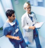 Picture of a female Physician walking in a Hospital hallway with a male Nurse. They are in talking and walking motion. The female Physician is holding a clipboard with paperwork on it and wearing a medical coat, scrubs, a mask hanging down around her neck, and a stethoscope around her neck.